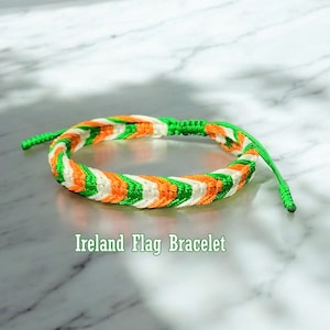 Pode incluir: Um bracelete de amizade verde, branco e laranja com um padrão de chevron. O bracelete é amarrado com um nó e tem uma cauda longa. O texto "Ireland Flag Bracelet" está escrito abaixo do bracelete.