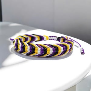 Puede incluir: Pulsera trenzada con un patrón de hilos morados, amarillos, negros y blancos. La pulsera está anudada y tiene extremos ajustables. La pulsera está sobre una superficie blanca.