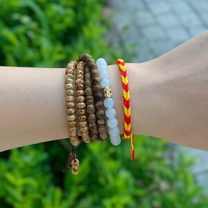 May include: A set of five bracelets stacked on a wrist. The bracelets are made of different materials, including wood beads, white stones, and a red and yellow braided cord.