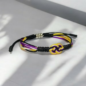 Könnte beinhalten: Ein geflochtenes Armband in Schwarz, Weiß, Gelb und Lila mit einem Knoten in der Mitte. Das Armband hat silberne Perlen.