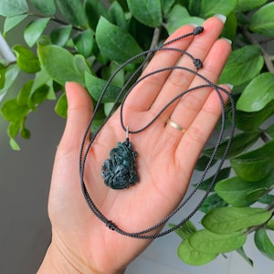 Puede incluir: Un colgante de jade verde oscuro tallado en forma de criatura mítica, colgado de un collar de cordón negro.