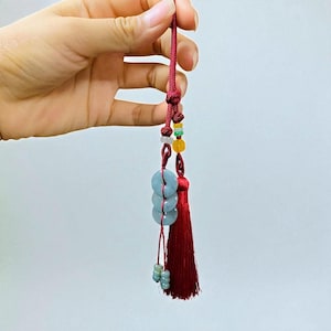 Puede incluir: Un amuleto de borla roja con un cordón anudado, adornado con cuentas de colores y tres colgantes circulares de color azul claro, similares al jade. El amuleto se sostiene sobre un fondo liso, mostrando su intrincado diseño y colores vibrantes.