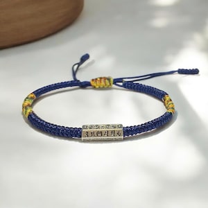 May include: A blue braided bracelet with a silver rectangular charm. The bracelet has yellow and green accents near the clasp. The charm has engraved characters. The bracelet is on a white surface.