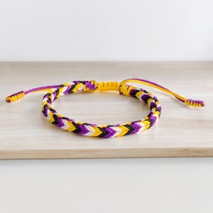 Puede incluir: Pulsera trenzada con un patrón de chevrón en morado, amarillo, negro y blanco. La pulsera tiene cordones ajustables en morado y amarillo. La pulsera se muestra sobre una superficie de madera clara.