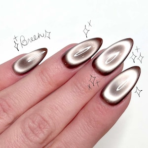 May include: A hand with long, oval-shaped nails painted with a brown and light brown cat-eye effect. The nails have a dark brown border and a shimmering, light brown center. The word "Breen" is written in cursive.