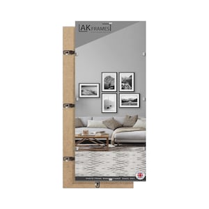 May include: A large rectangular mirror with a light brown wooden frame and metal fixings. The mirror reflects a living room with framed black and white photographs, a sofa, and a patterned rug. The text "AK Frames" is visible.
