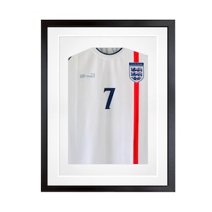 May include: A white England football shirt featuring the number 7 in dark blue, a vertical red stripe, and the England team crest, presented in a black frame with a white mount. The text AK FRAMES is printed on the right side of the jersey chest area.