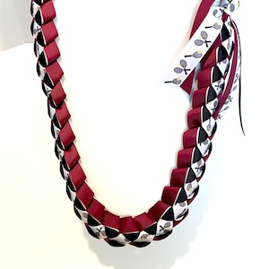 Puede incluir: Un lei de cinta trenzada en burdeos, negro y blanco. El lei presenta un patrón repetitivo de cintas burdeos, intercaladas con cintas negras y blancas. Una sección del lei tiene una cinta blanca con gráficos de raquetas de tenis.