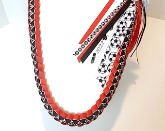 May include: Red, black, and white soccer themed lei with a graduation cap and the year 2022 on a white ribbon.