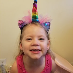 Pode incluir: Uma menina pequena usando uma faixa de cabeça de unicórnio rosa e arco-íris com um chifre e orelhas brilhantes. Ela está sorrindo e olhando para a câmera.