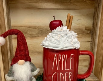 May include: A red ceramic mug with "APPLE CIDER" in white lettering, topped with whipped cream, an apple, and cinnamon sticks. A gnome with a red hat and striped legs sits beside the mug. The items are displayed on a wooden shelf.