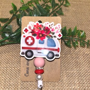 May include: A white and red ambulance badge reel with a floral design. The ambulance has a red cross and is decorated with red and pink flowers. The reel has a pink bead, red beads, and a clear, glittery charm.
