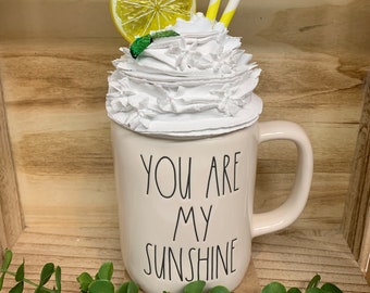 May include: Cream-colored mug with the text "YOU ARE MY SUNSHINE" in black. The mug is topped with white whipped cream, a lemon slice, and yellow and white straws. A decorative item.