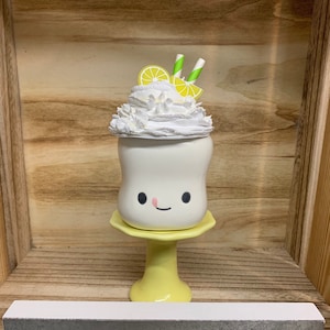 May include: A whimsical, white ceramic milkshake with a smiling face, topped with whipped cream, lemon slices, and striped straws. It sits on a yellow pedestal. The sign below reads "Easy Peasy Lemon Squeezy".