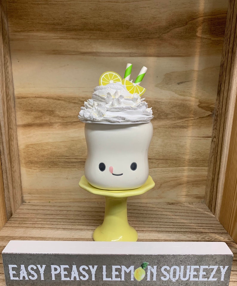May include: A whimsical, white ceramic milkshake with a smiling face, topped with whipped cream, lemon slices, and striped straws. It sits on a yellow pedestal. The sign below reads "Easy Peasy Lemon Squeezy".