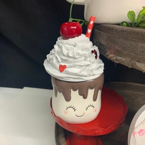May include: A whimsical ceramic mug shaped like a marshmallow, topped with white whipped cream, a red cherry, and a red straw. The mug has a smiling face and sits on a red pedestal. Decorative item for Valentine's Day.