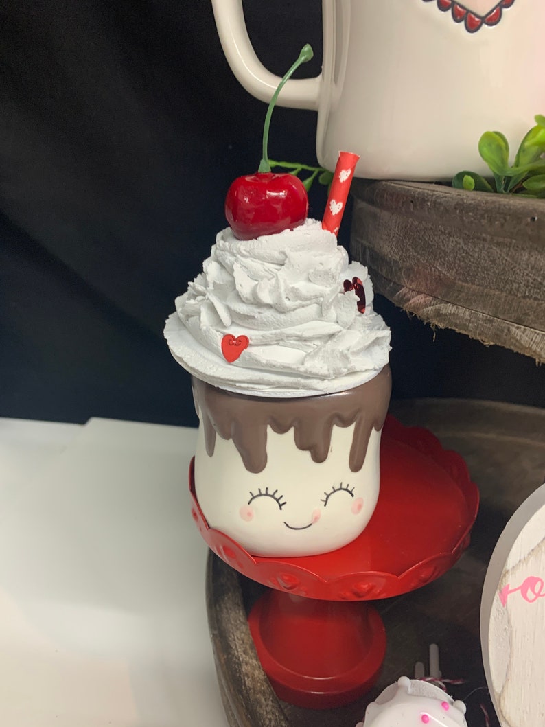 May include: A whimsical ceramic mug shaped like a marshmallow, topped with white whipped cream, a red cherry, and a red straw. The mug has a smiling face and sits on a red pedestal. Decorative item for Valentine's Day.
