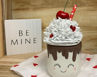 May include: A Valentine's Day-themed ceramic mug with a smiling face, topped with white whipped cream, a red cherry, and a red straw. The mug has a chocolate-colored rim and is decorated with small red hearts. A wooden sign with the words "BE MINE" is in the background.