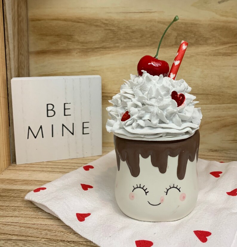 May include: A Valentine's Day-themed ceramic mug with a smiling face, topped with white whipped cream, a red cherry, and a red straw. The mug has a chocolate-colored rim and is decorated with small red hearts. A wooden sign with the words "BE MINE" is in the background.