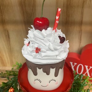May include: A decorative Valentine's Day themed mug with a smiling face, topped with white whipped cream, a red cherry, and a straw. The mug sits on a red pedestal and is decorated with chocolate drips.