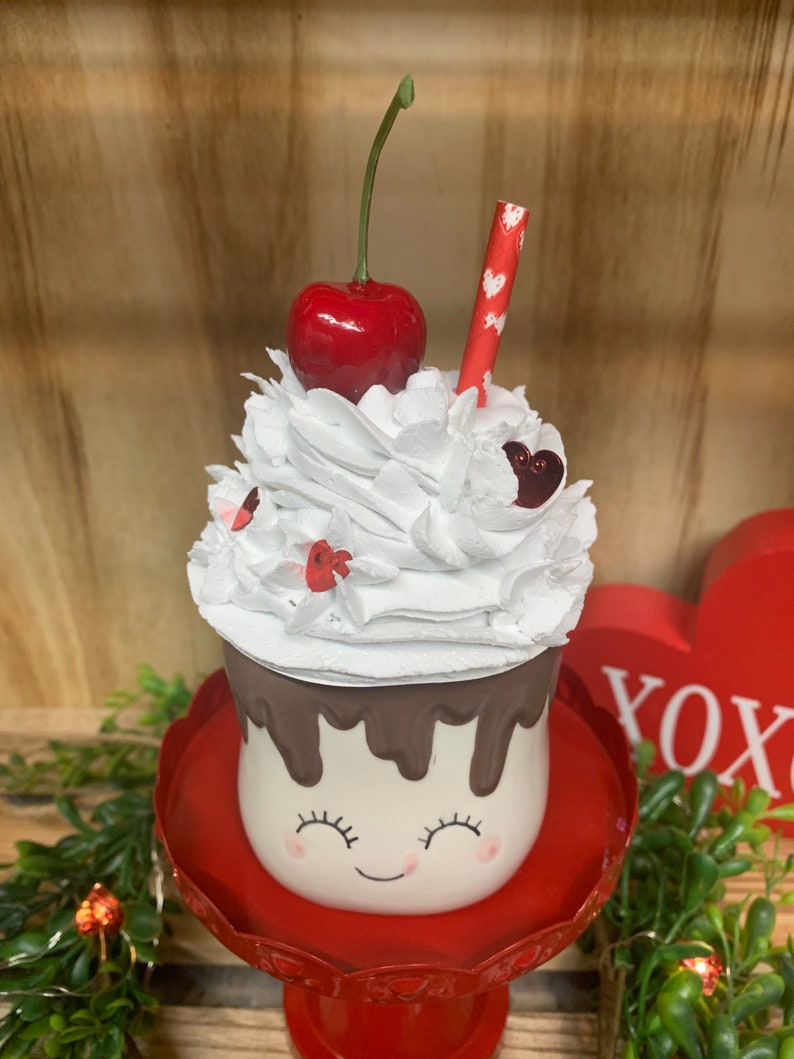 May include: A decorative Valentine's Day themed mug with a smiling face, topped with white whipped cream, a red cherry, and a straw. The mug sits on a red pedestal and is decorated with chocolate drips.