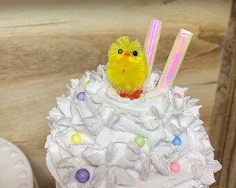 May include: A decorative white ceramic cup topped with white frosting, colorful sprinkles, and a yellow chick. Two iridescent straws are placed in the frosting. The cup is set against a light brown wooden background.