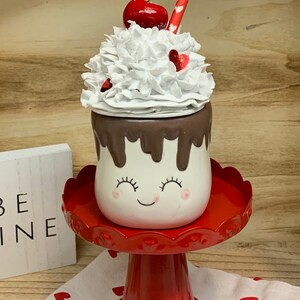 May include: A whimsical ceramic marshmallow-shaped container with a smiling face, chocolate drizzle, and whipped cream topping, adorned with a cherry and a red straw. It sits on a red pedestal, with a small sign that says "BE MINE".