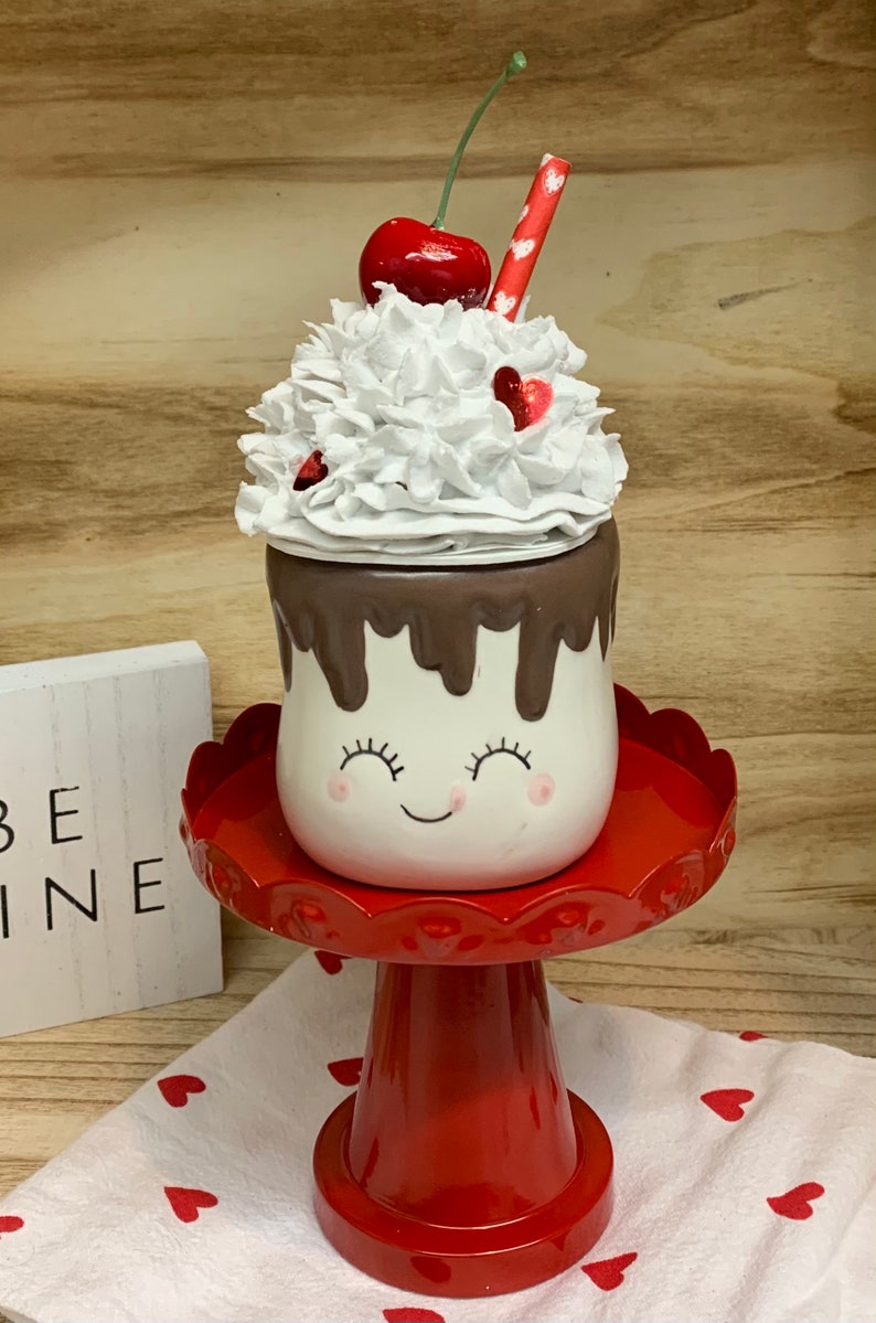 May include: A whimsical ceramic marshmallow-shaped container with a smiling face, chocolate drizzle, and whipped cream topping, adorned with a cherry and a red straw. It sits on a red pedestal, with a small sign that says "BE MINE".
