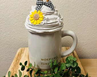 May include: A decorative coffee mug with a white whipped cream topping, adorned with a yellow sunflower, a black and white checkered bow, and two cinnamon sticks. The mug has the words "MORNING SUNSHINE" printed on it.
