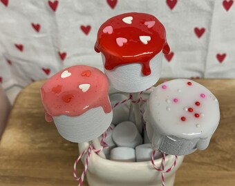 May include: A ceramic container with a bunny face holds marshmallow pops. The pops have white marshmallow bases and are topped with red, pink, and white icing decorated with heart sprinkles. The container is tied with pink and white twine.