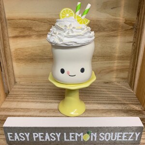 May include: A whimsical, white ceramic drink with a smiling face, topped with whipped cream, lemon slices, and straws. It sits on a yellow pedestal and a sign below reads "EASY PEASY LEMON SQUEEZY".