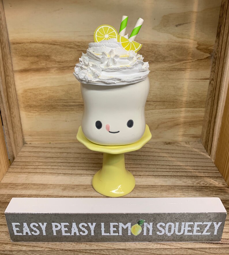 May include: A whimsical, white ceramic drink with a smiling face, topped with whipped cream, lemon slices, and straws. It sits on a yellow pedestal and a sign below reads "EASY PEASY LEMON SQUEEZY".