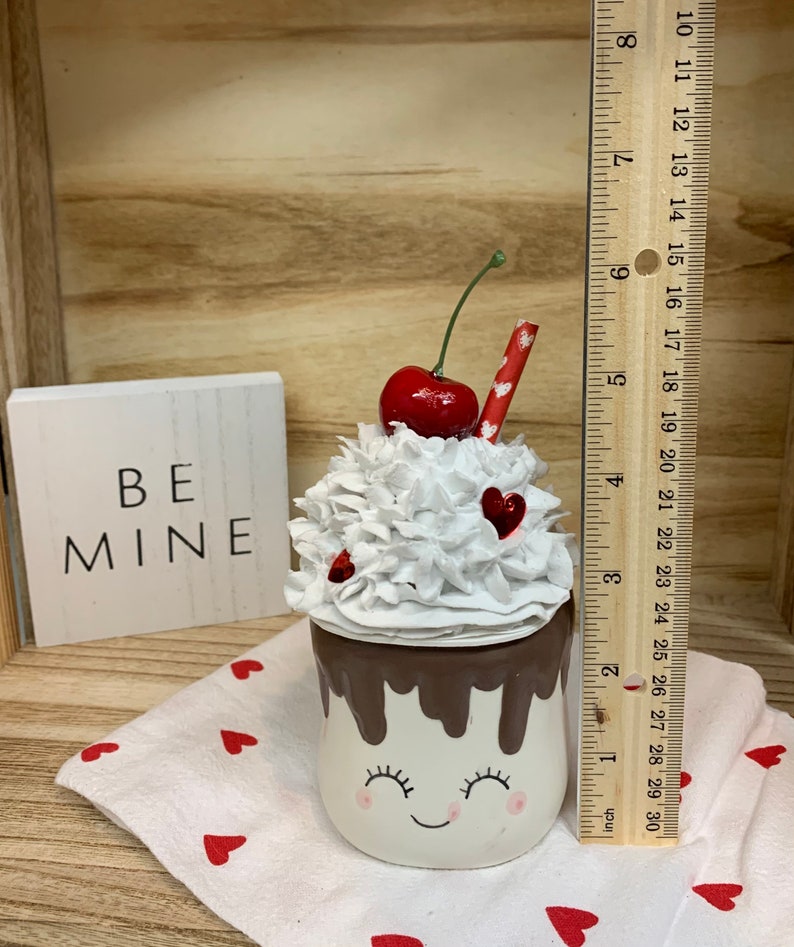 May include: A decorative Valentine's Day themed ceramic mug, approximately 3 inches tall, with a smiling face, whipped cream topping, a cherry, and a red straw. The mug is decorated with red hearts and the words "BE MINE" on a small white block.