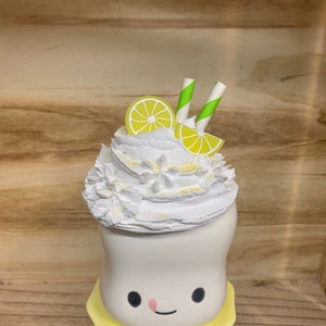 May include: A whimsical, white ceramic dessert figurine with a smiling face, topped with whipped cream, lemon slices, and green-striped straws. The figurine sits on a yellow base, creating a cheerful, summery vibe. The dessert is decorated with yellow glitter.
