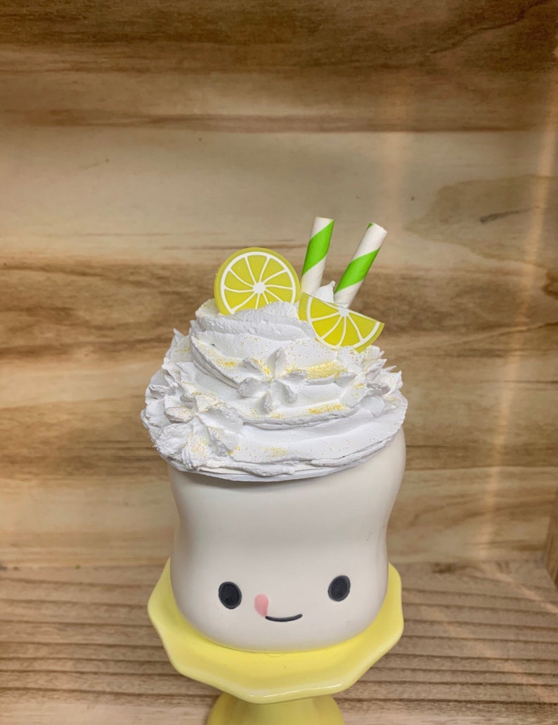 May include: A whimsical, white ceramic dessert figurine with a smiling face, topped with whipped cream, lemon slices, and green-striped straws. The figurine sits on a yellow base, creating a cheerful, summery vibe. The dessert is decorated with yellow glitter.