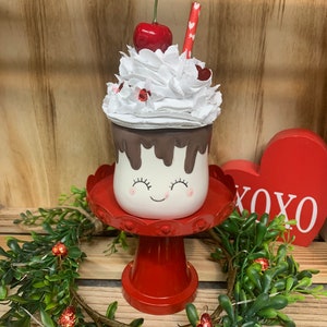 May include: A Valentine's Day-themed decorative ceramic milkshake with a smiling face, chocolate drips, and whipped cream topped with a cherry and a straw. It sits on a red pedestal with a heart-shaped XOXO sign in the background.