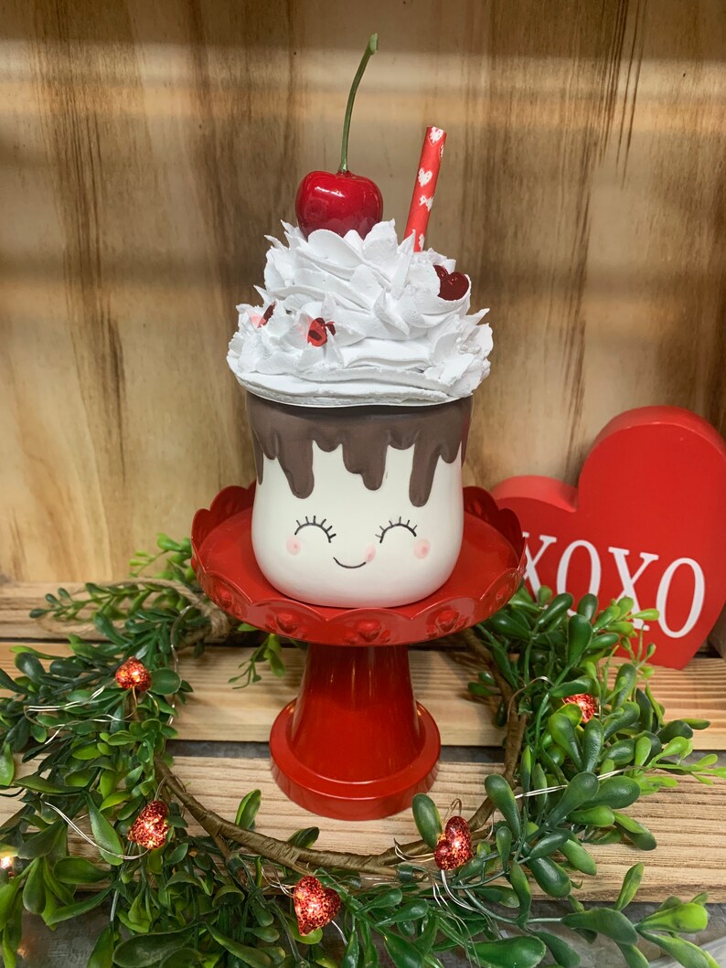 May include: A Valentine's Day-themed decorative ceramic milkshake with a smiling face, chocolate drips, and whipped cream topped with a cherry and a straw. It sits on a red pedestal with a heart-shaped XOXO sign in the background.
