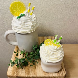 May include: Two decorative faux lemon drinks. One is in a gray mug and the other in a white cup. Both have white whipped cream, lemon slices, yellow and green striped straws, and a sprig of mint. They sit on a wooden tray with greenery.