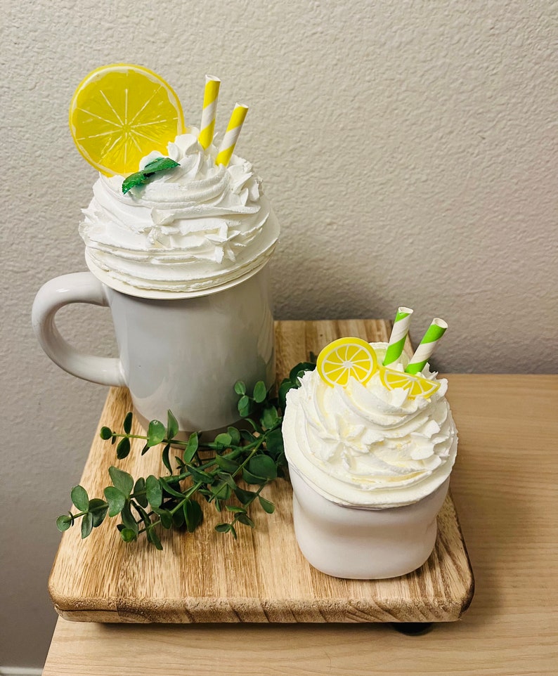 May include: Two decorative faux lemon drinks. One is in a gray mug and the other in a white cup. Both have white whipped cream, lemon slices, yellow and green striped straws, and a sprig of mint. They sit on a wooden tray with greenery.