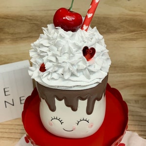 May include: A decorative ceramic dessert cup with a smiling face, topped with white whipped cream, a red cherry, and a heart-shaped accent. The cup sits on a red pedestal, creating a charming, whimsical design.