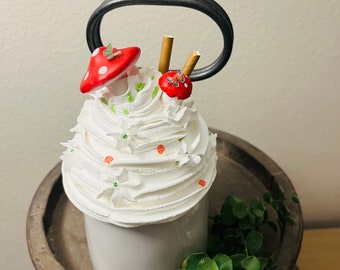 May include: A decorative white ceramic mug topped with whipped cream and whimsical mushroom and berry accents. The mug has a black handle and sits on a wooden tiered tray. Greenery adds a natural touch.