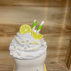 May include: A whimsical, white ceramic cup with a smiling face, topped with whipped cream, lemon slices, and green and white striped straws. The cup sits on a yellow saucer, creating a playful, summery aesthetic.