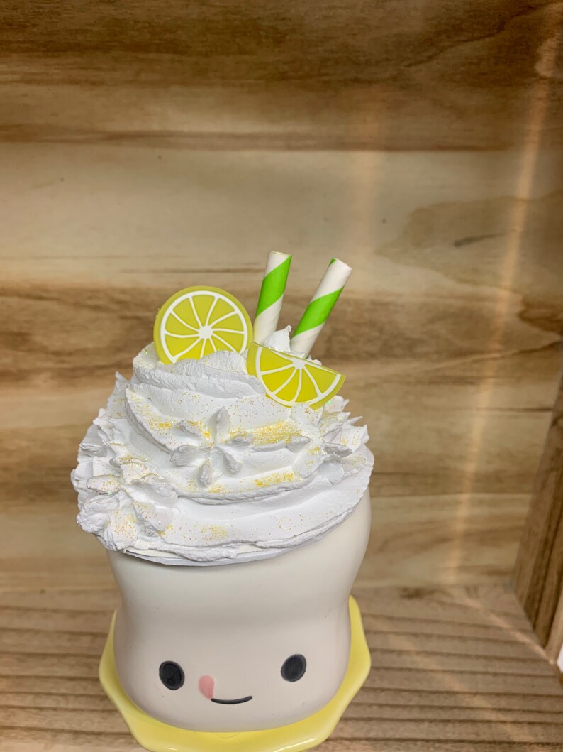 May include: A whimsical, white ceramic cup with a smiling face, topped with whipped cream, lemon slices, and green and white striped straws. The cup sits on a yellow saucer, creating a playful, summery aesthetic.
