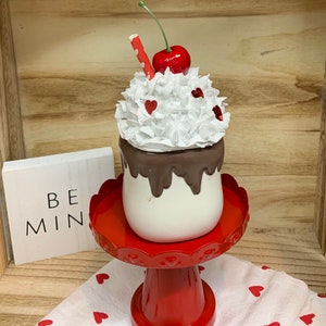 May include: A decorative Valentine's Day milkshake with white whipped cream, a red cherry, and a red and white straw. The milkshake sits on a red pedestal stand with a scalloped edge. A small sign reads "BE MIN".