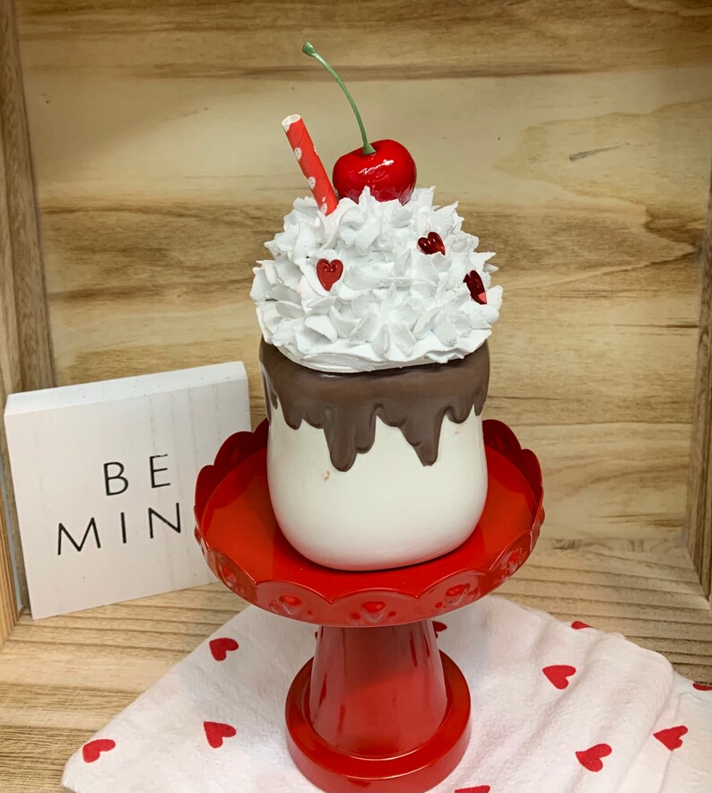 May include: A decorative Valentine's Day milkshake with white whipped cream, a red cherry, and a red and white straw. The milkshake sits on a red pedestal stand with a scalloped edge. A small sign reads "BE MIN".
