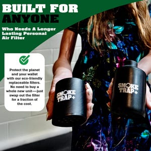 May include: Two black cylindrical air purifiers with the text "Smoke Trap+" in white. The purifiers are being held by a person wearing a floral dress.