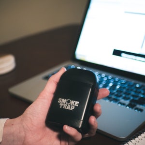 May include: A black, rectangular device with the text "SMOKE TRAP" in white lettering. The device is being held in a person's hand.