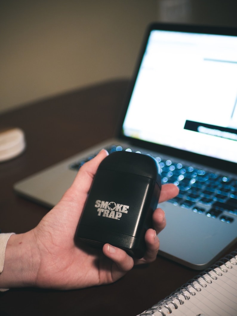 May include: A black, rectangular device with the text "SMOKE TRAP" in white lettering. The device is being held in a person's hand.