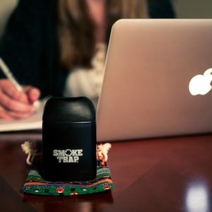 May include: A black cylindrical container with the text "Smoke Trap" printed on it. The container is sitting on a colorful patterned fabric pouch. A silver laptop computer with an Apple logo is in the background.