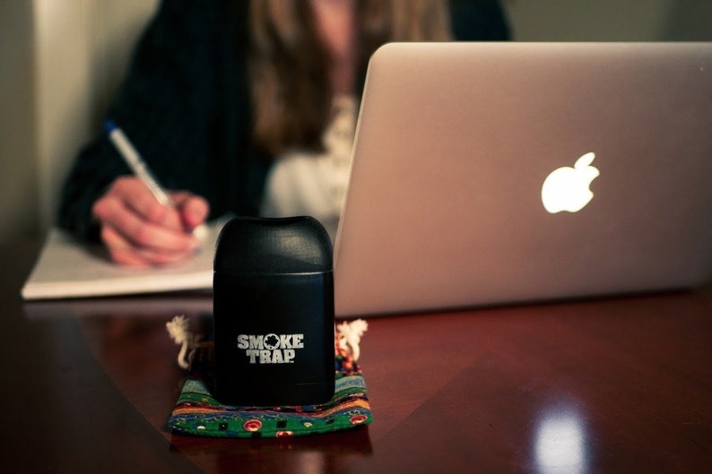 May include: A black cylindrical container with the text "Smoke Trap" printed on it. The container is sitting on a colorful patterned fabric pouch. A silver laptop computer with an Apple logo is in the background.